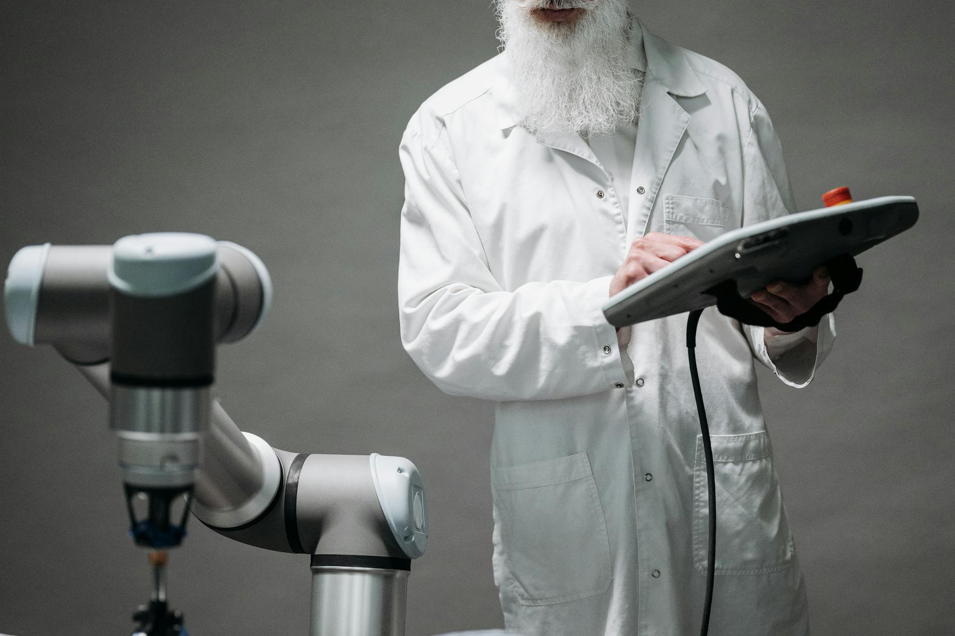 Scientist in a white lab coat uses a tablet to control a robotic arm in a lab setting with gray background