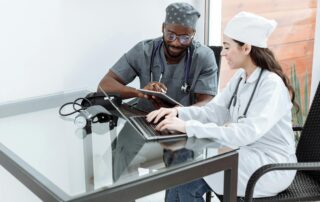 Two healthcare professionals review notes on a tablet and laptop at a glass desk.