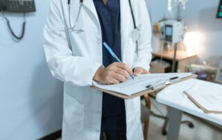 Doctor in a white coat signs a clipboard form with a blue pen in a clinical room, stethoscope around the neck.