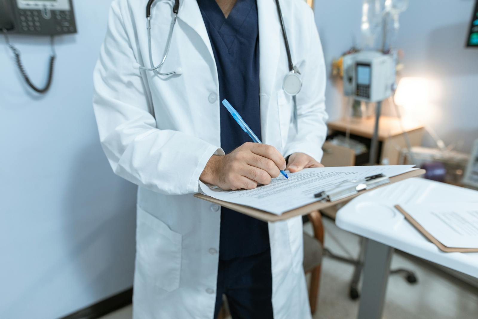 Doctor in a white coat signs a clipboard form with a blue pen in a clinical room, stethoscope around the neck.
