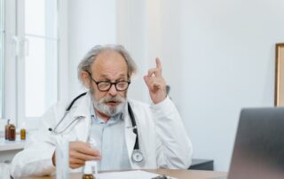 Older doctor in a white coat with a stethoscope around his neck sits at a desk, raising a finger as he speaks. (informative)