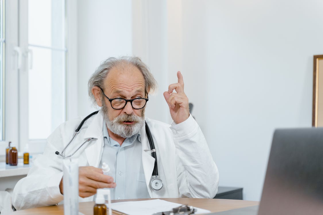 Older doctor in a white coat with a stethoscope around his neck sits at a desk, raising a finger as he speaks. (informative)