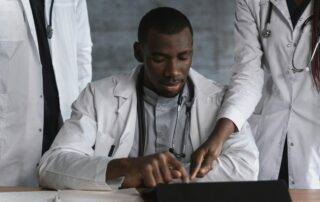 Group of doctors in white coats gathered around a table, examining a tablet and documents together at a desk with stethoscopes nearby.