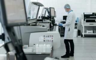 Lab technician in a mask and hairnet operates automated medical testing machines in a clean, modern laboratory.
