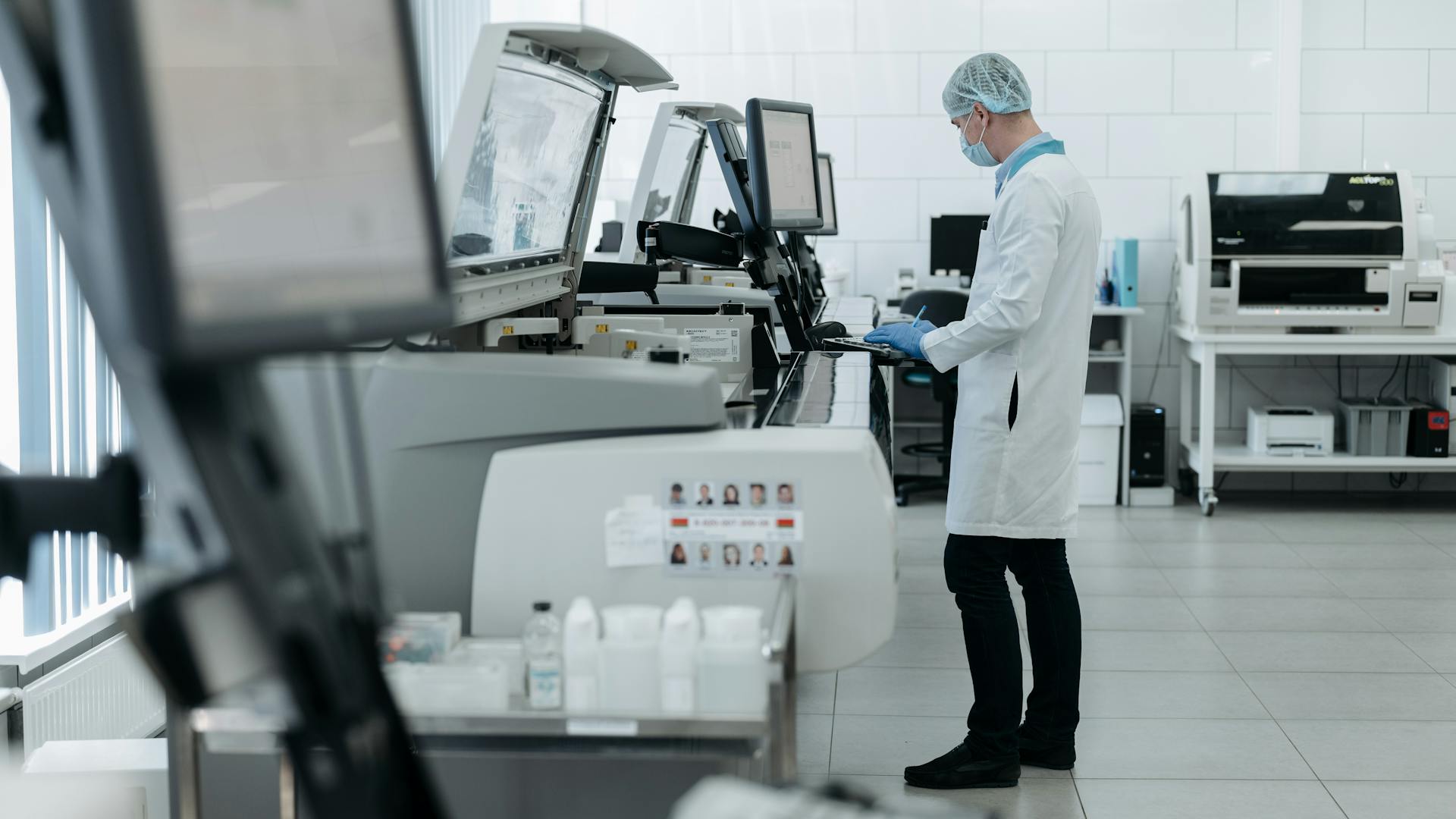 Lab technician in a mask and hairnet operates automated medical testing machines in a clean, modern laboratory.