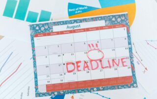 August calendar page with a bold red 'DEADLINE' stamp across the days and a circled date around the 13th, surrounded by charts and papers.