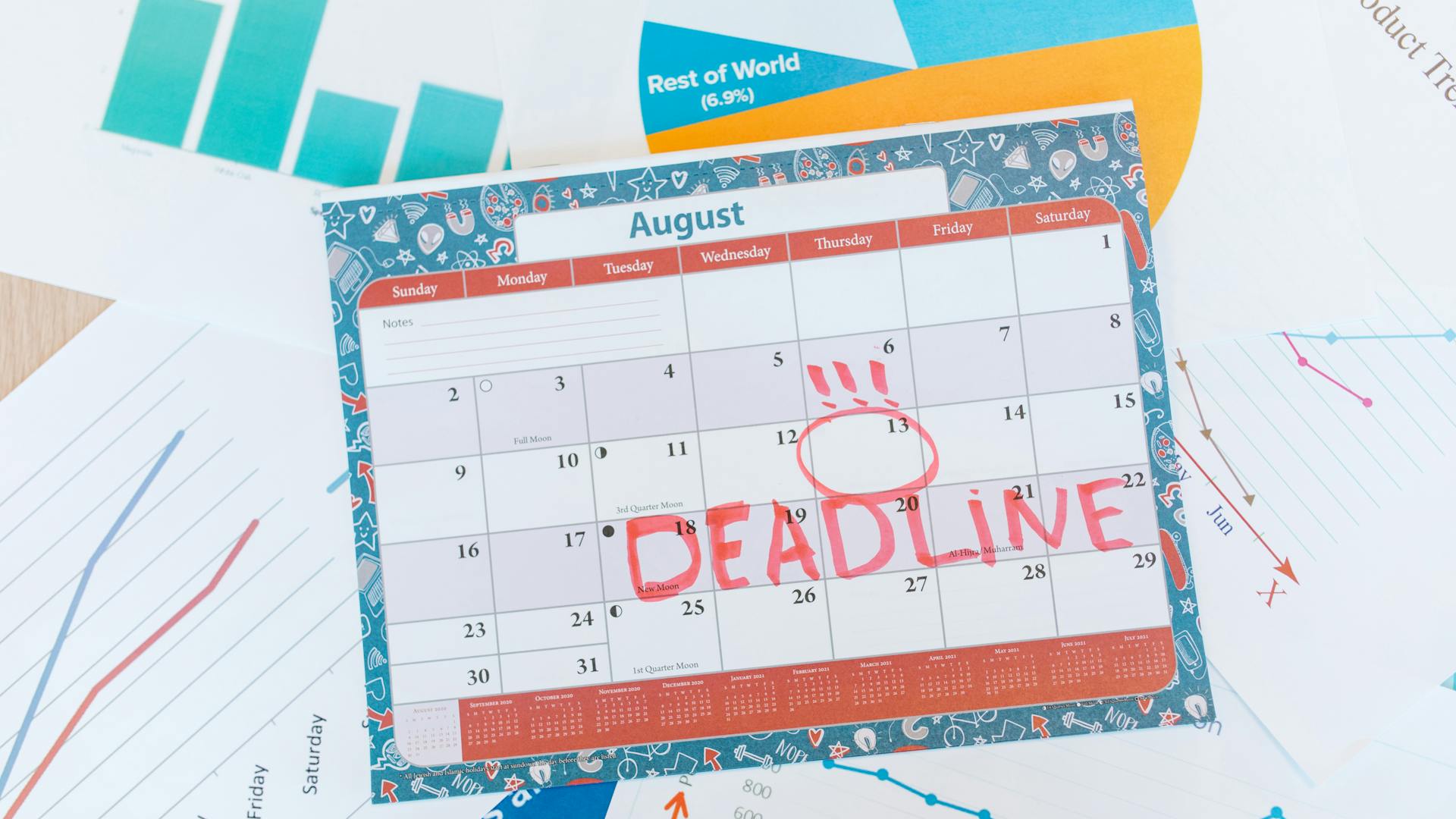 August calendar page with a bold red 'DEADLINE' stamp across the days and a circled date around the 13th, surrounded by charts and papers.