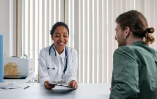 Female doctor in a white coat with stethoscope consulting a patient across a desk, tablet in hand.