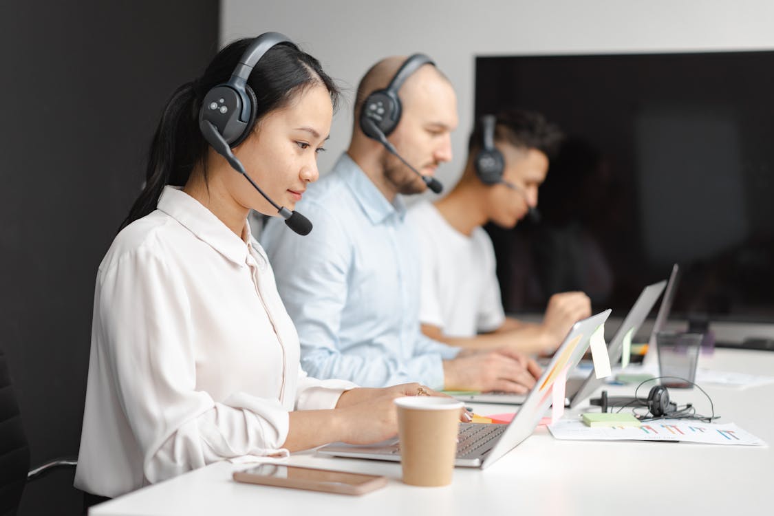 Three customer service agents wearing headsets and working on laptops at a shared desk in a call-center setup.