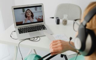 Person wearing a headset at a white desk, watching a video call on a laptop with notepaper and a pill bottle nearby.