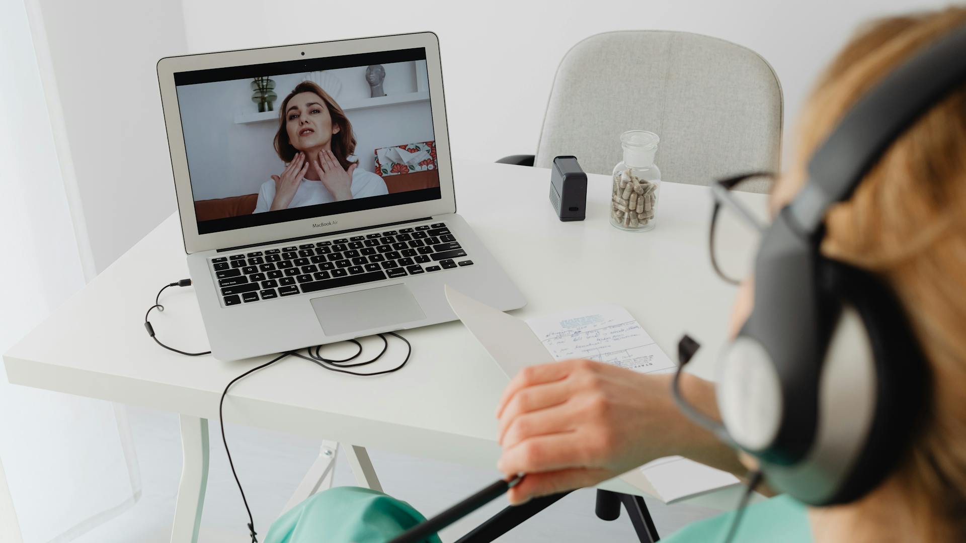 Person wearing a headset at a white desk, watching a video call on a laptop with notepaper and a pill bottle nearby.