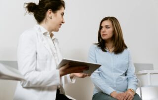 Doctor in a white coat shows a tablet to a seated patient in a light blue shirt during a consultation in a clinical setting