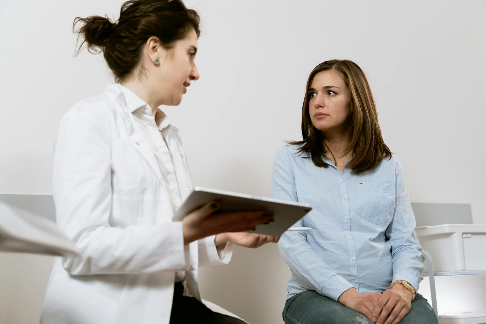 Doctor in a white coat shows a tablet to a seated patient in a light blue shirt during a consultation in a clinical setting