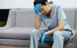 Medical professional in light blue scrubs and blue gloves sitting on a gray couch, head resting on hand in a stressed pose, wearing a mask pulled down below the chin.