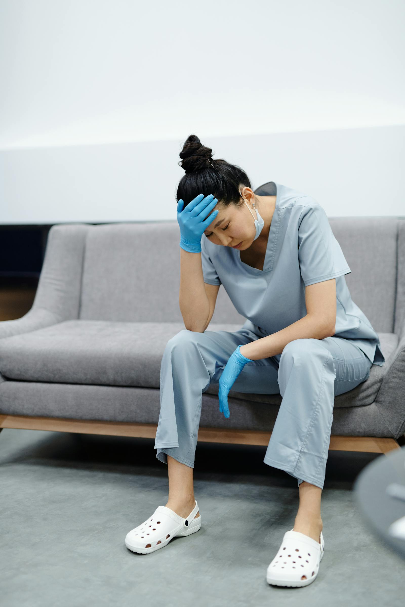 Medical professional in light blue scrubs and blue gloves sitting on a gray couch, head resting on hand in a stressed pose, wearing a mask pulled down below the chin.