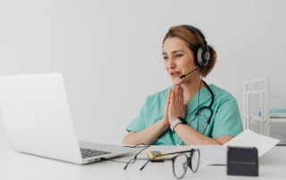 Healthcare professional in teal scrubs with a headset at a laptop, hands pressed together in a prayer-like gesture.