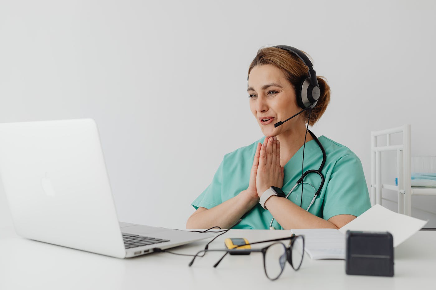 Healthcare professional in teal scrubs with a headset at a laptop, hands pressed together in a prayer-like gesture.