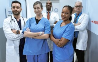 Team of six healthcare professionals in blue scrubs and white coats posing in a hospital corridor.