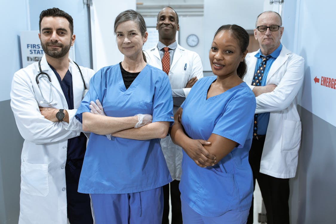 Team of six healthcare professionals in blue scrubs and white coats posing in a hospital corridor.