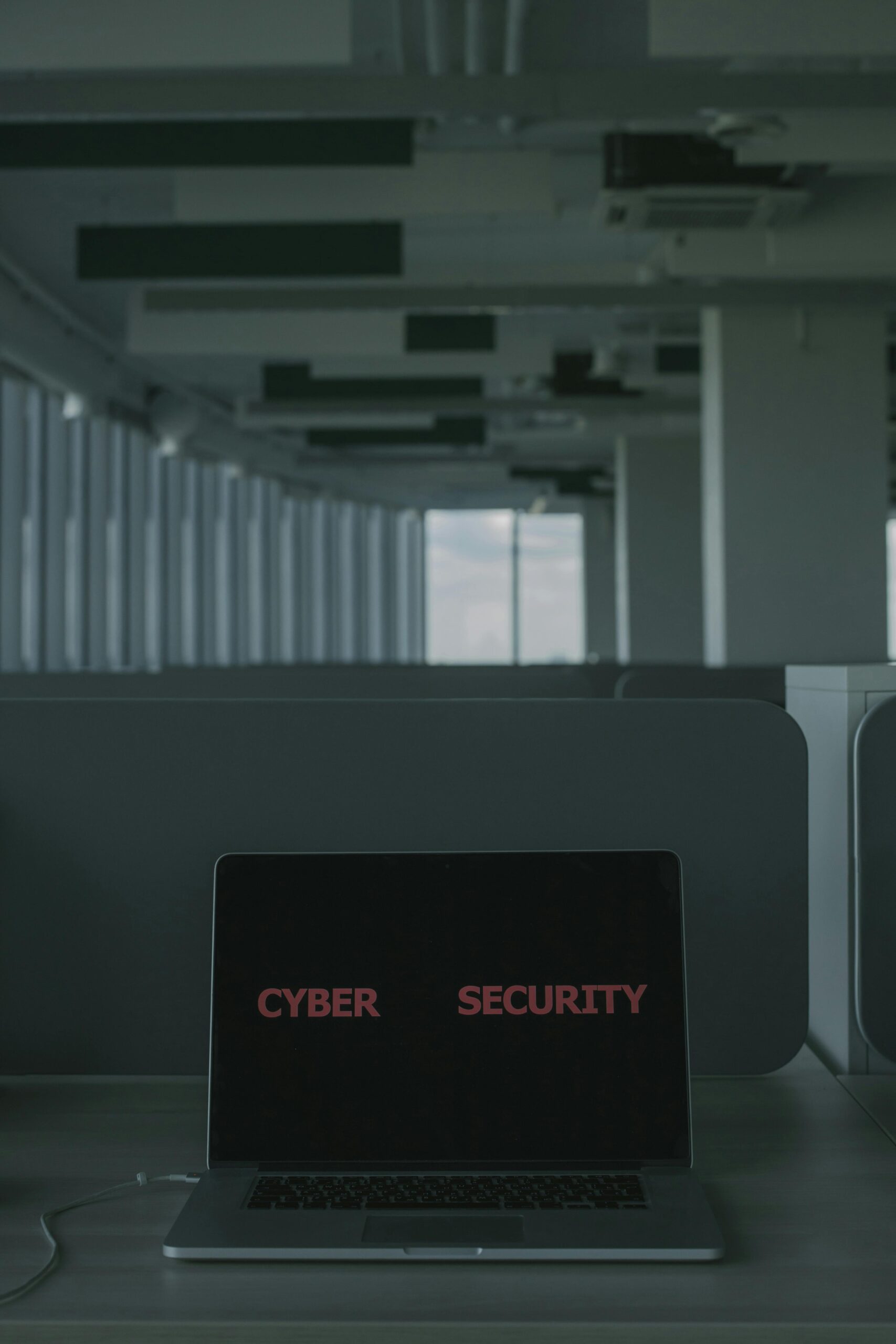 Laptop on desk in a dim office; screen shows the words 'CYBER SECURITY' in red font.
