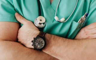 Crossed arms of a clinician in teal scrubs with a stethoscope around the neck and pens in the pocket