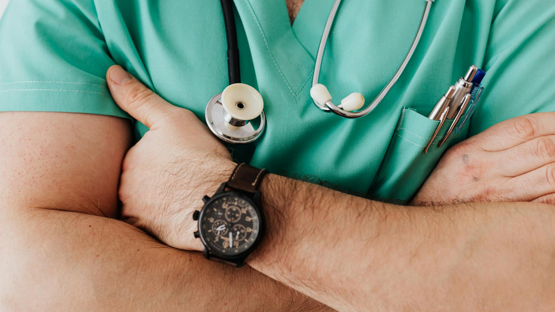 Crossed arms of a clinician in teal scrubs with a stethoscope around the neck and pens in the pocket