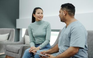 Asian woman in a mint-green turtleneck smiles as she talks to a man in light-blue scrubs holding a tablet on a gray sofa in a living room.