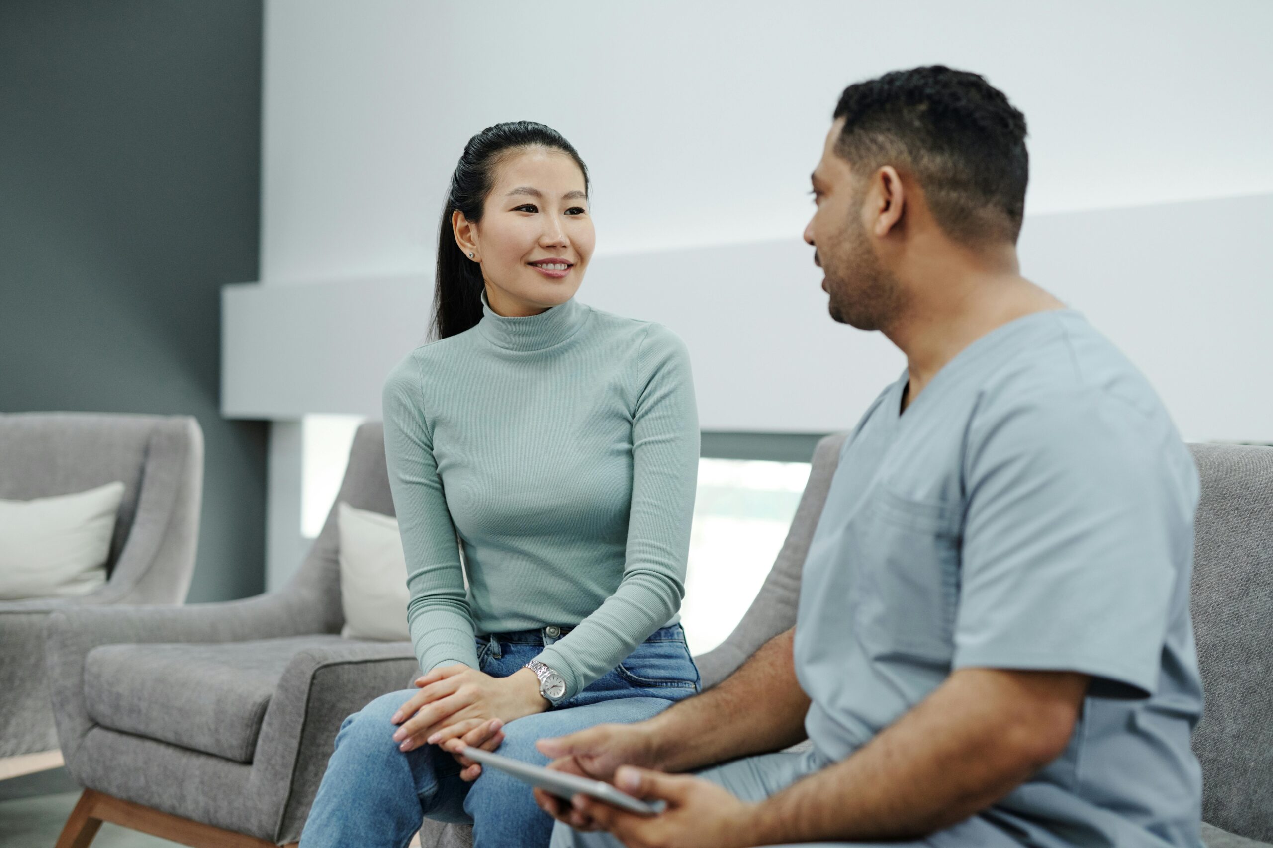 Asian woman in a mint-green turtleneck smiles as she talks to a man in light-blue scrubs holding a tablet on a gray sofa in a living room.