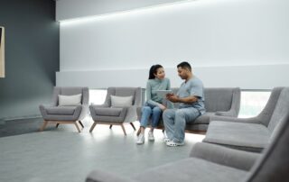 A man in scrubs and a woman in casual clothes sit on gray lounge chairs in a modern waiting area, looking at a tablet together.