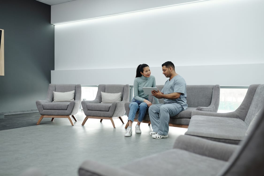 A man in scrubs and a woman in casual clothes sit on gray lounge chairs in a modern waiting area, looking at a tablet together.