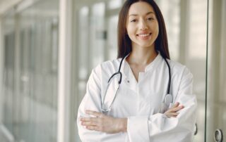 Female doctor in a white coat with a stethoscope around her neck, smiling, arms crossed in a clinical hallway