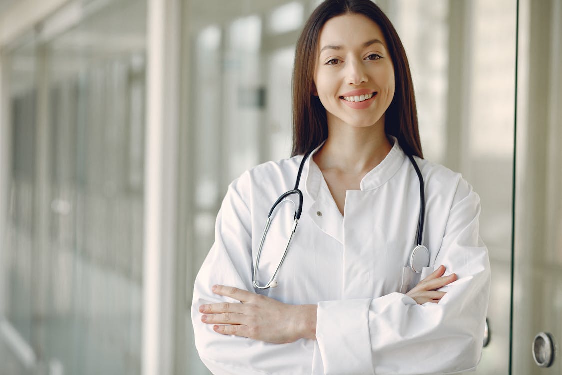 Female doctor in a white coat with a stethoscope around her neck, smiling, arms crossed in a clinical hallway