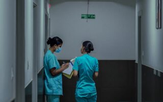 Two nurses in teal scrubs in a hospital hallway, one wearing a mask and writing on a clipboard.