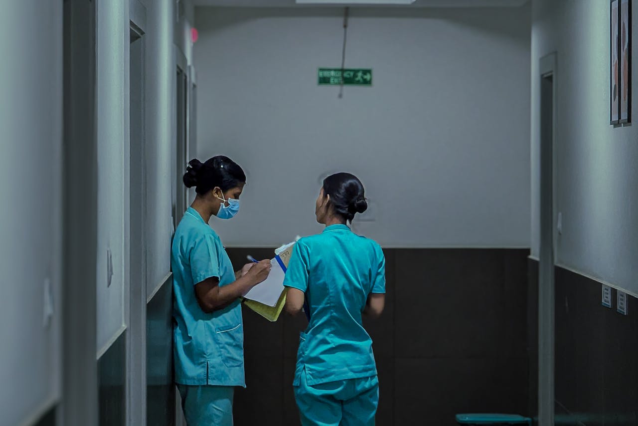 Two nurses in teal scrubs in a hospital hallway, one wearing a mask and writing on a clipboard.