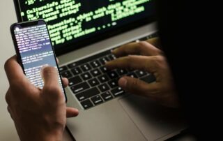 Person types code on a laptop keyboard while a smartphone in the foreground also displays lines of code on its screen.