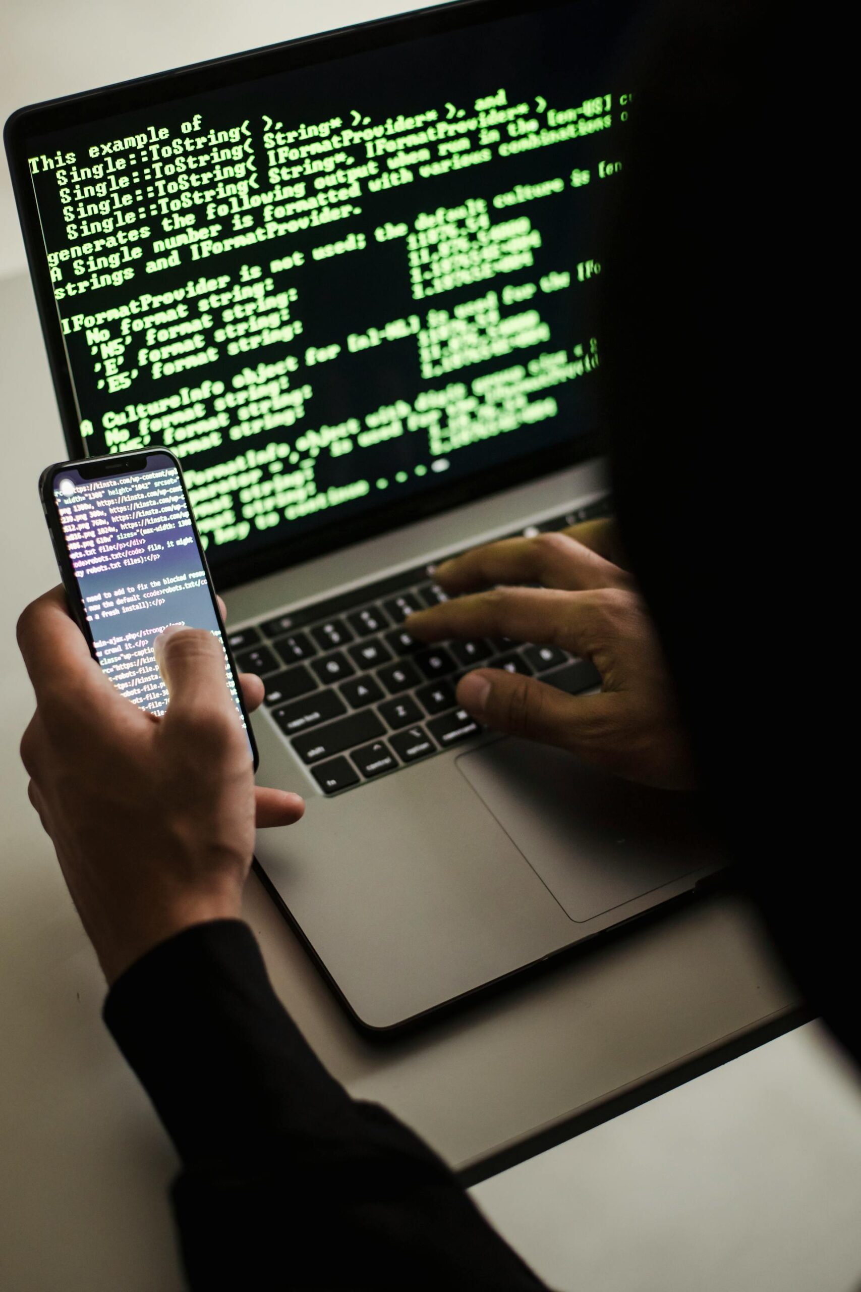 Person types code on a laptop keyboard while a smartphone in the foreground also displays lines of code on its screen.