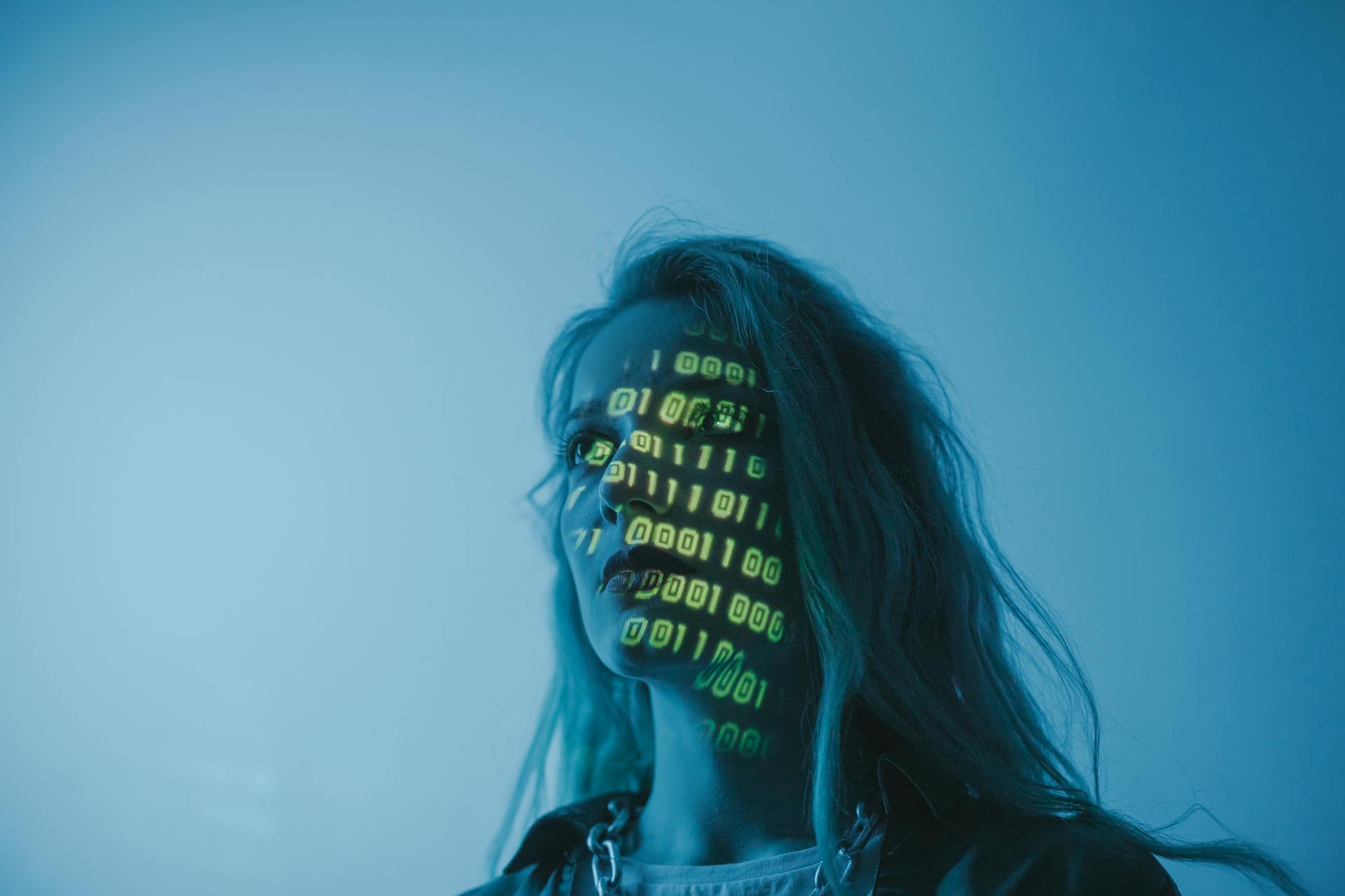 Side profile of a person with glowing binary code projected on their face against a blue background, suggesting digital identity or cybersecurity.