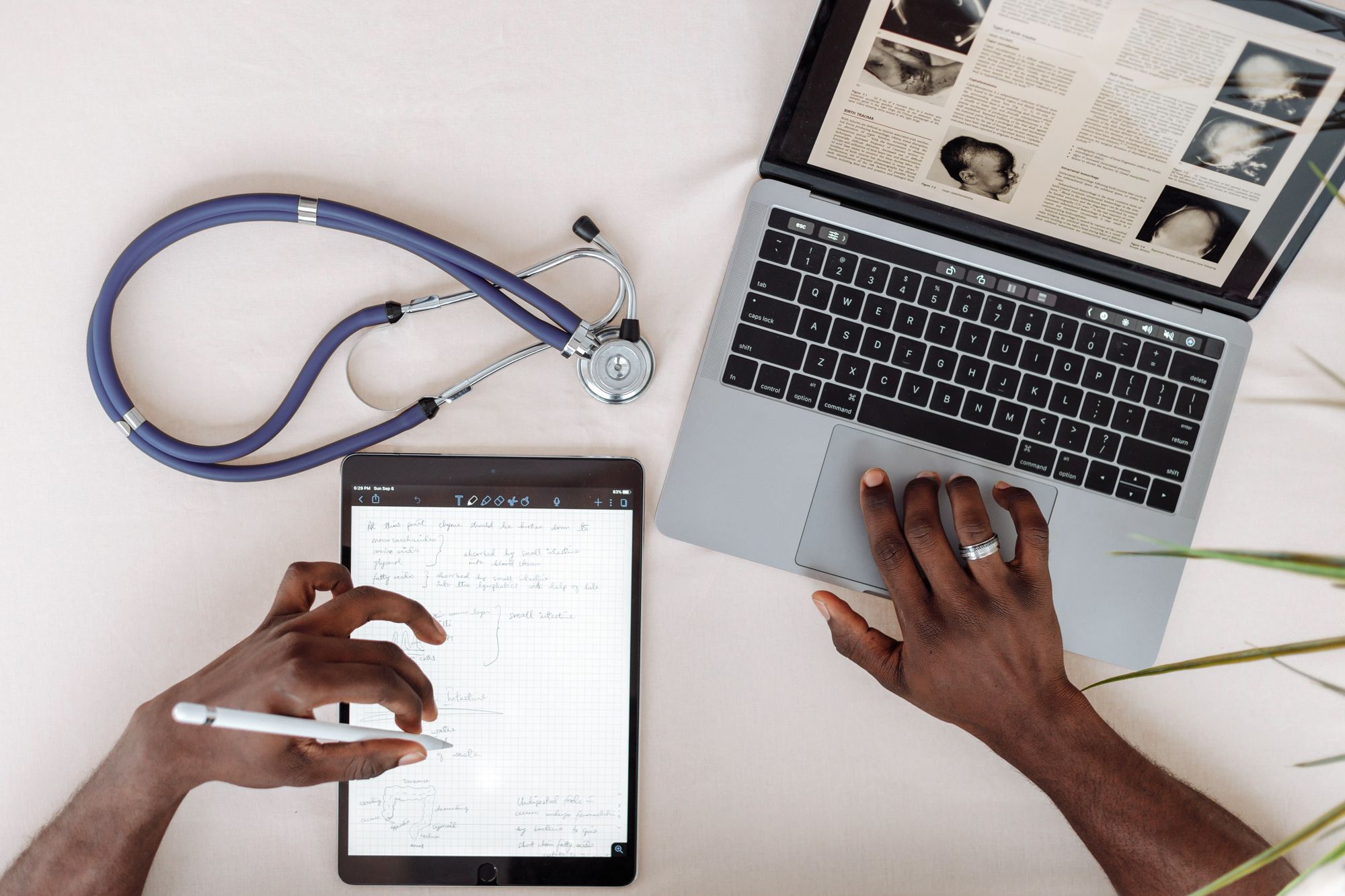 Clinician writing clinical notes by hand on a tablet beside a laptop and a stethoscope