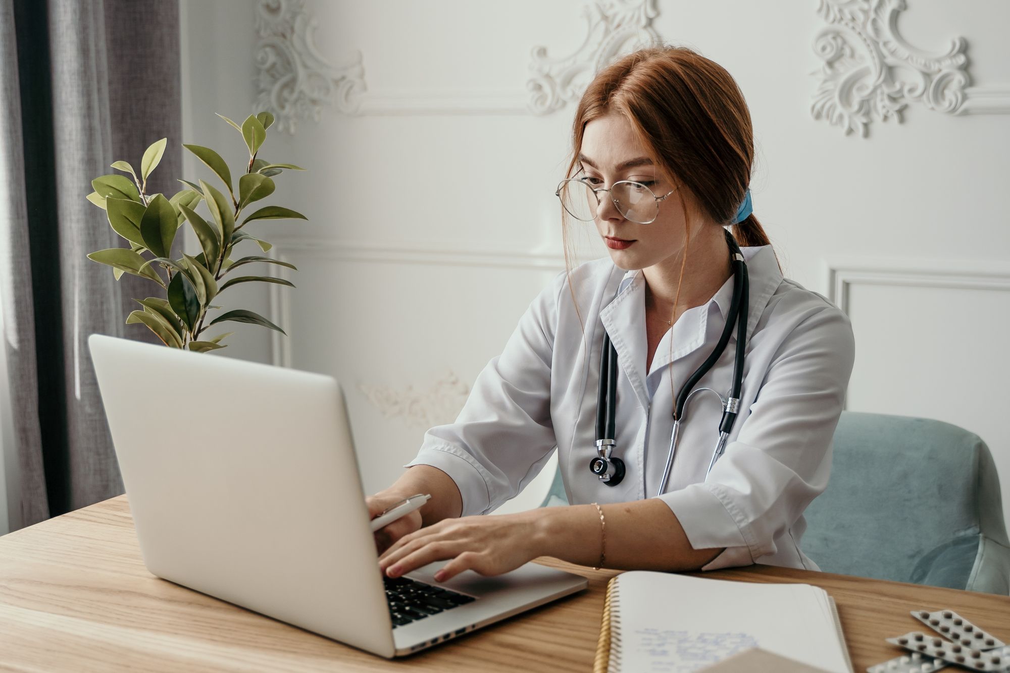 A clinician in a white coat working at a laptop in a healthcare office