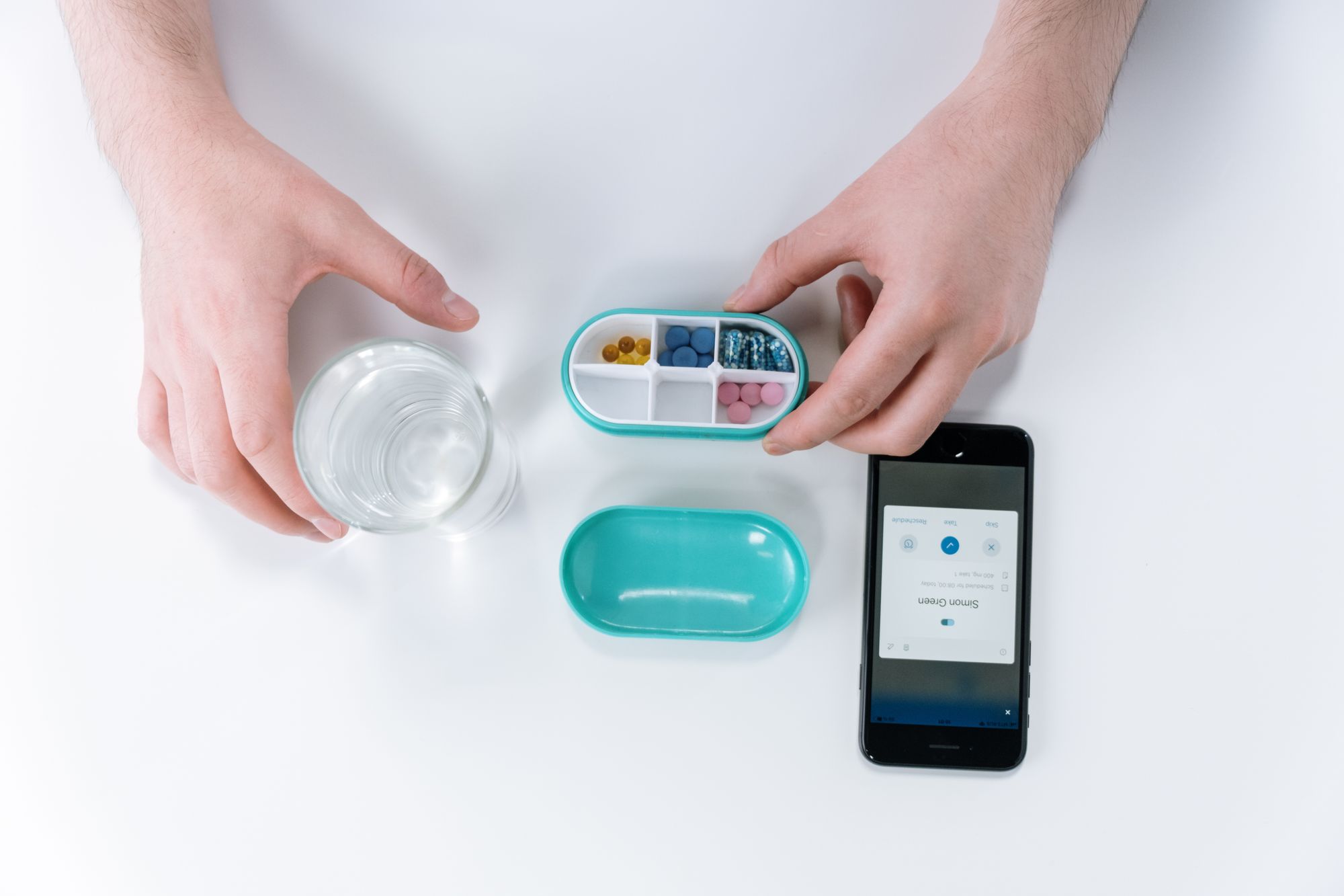 Hands with a weekly pill organizer, a glass of water and a phone showing a medication reminder