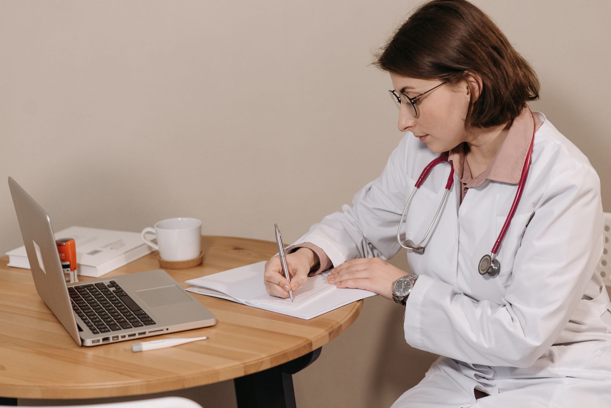 A physician writing notes at a desk with a laptop